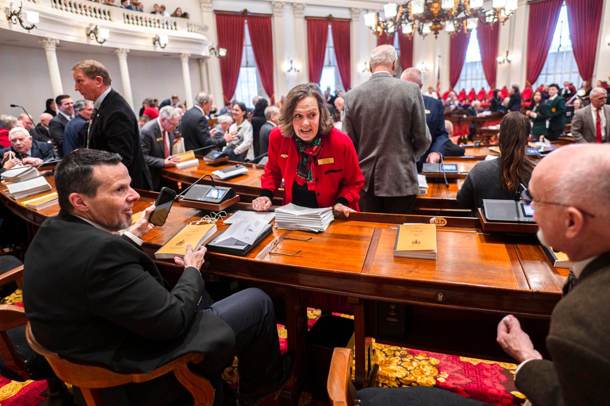 People converse and prepare materials in a large, formal legislative chamber with red curtains and wooden desks.