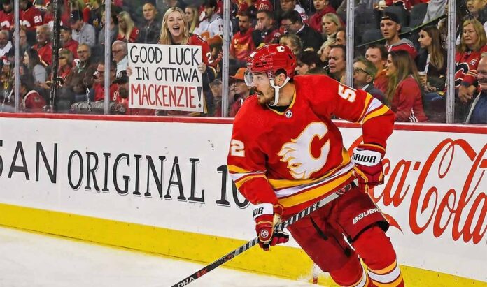 Calgary Flames defenseman MacKenzie Weegar skating with the puck along the boards during a game. Behind the glass, a smiling blonde fan holds up a handwritten white sign that reads, "GOOD LUCK IN OTTAWA MACKENZIE!", referencing trade rumors connecting him to the Ottawa Senators.