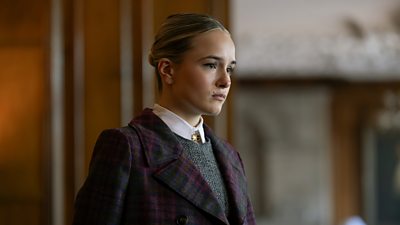 Photo of Miriam Petche in character as Sweetpea Golighty. Wearing a wool blazer, a grey jumper and white collared shirt, she looks soberly at something offscreen. 