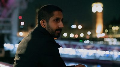 Photo of Sagar Radia in character as Rishi Ramdani. Wearing a black coat, he stands outside and leans on a railing in Central London. A blurred silhouette of Big Ben and the Houses of Parliament are visible in the background. 