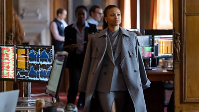 Photo of Myha’la in character as Harper. Wearing a grey overcoat, layered on top of a grey skirt-suit, she walks through an office. Computer screens on nearby desks show a variety of graphs and spreadsheets. 