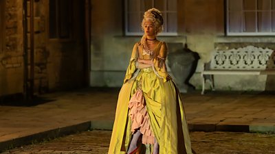 Photo of Marisa Abela in character as Yasmin Kara-Hanani. Wearing an elaborate, yellow and pink period gown, she stands outside with her arms folded. 