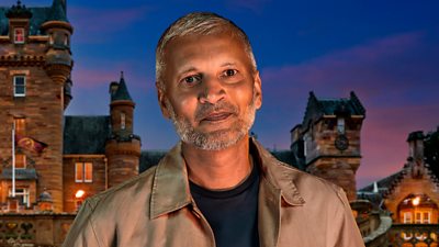 A man in a dark t-shirt and light brown shirt smiles to camera, set against the backdrop of a castle by night