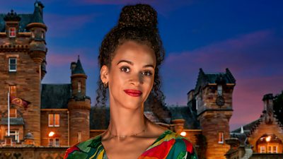 A woman in a multicoloured top smiles to camera wearing bold red lipstick, set against the background of a castle by night