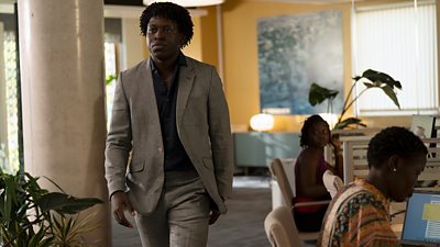A man wearing a grey suit over a blue shirt walks through an office. Plants decorate the space, alongside marble columns. In the background, two people work at desks. One watches as the man walks past. 