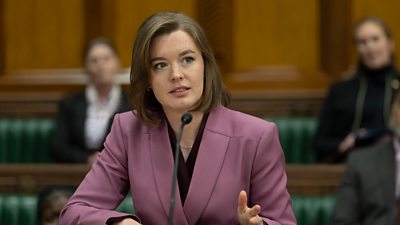 A woman wearing a pink blazer sits at a table with a microphone. In the background, people are sat on benches with green fabric. 