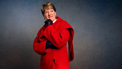 A woman in a red coat stands with one hand to her chin, smiling as she looks at something off screen