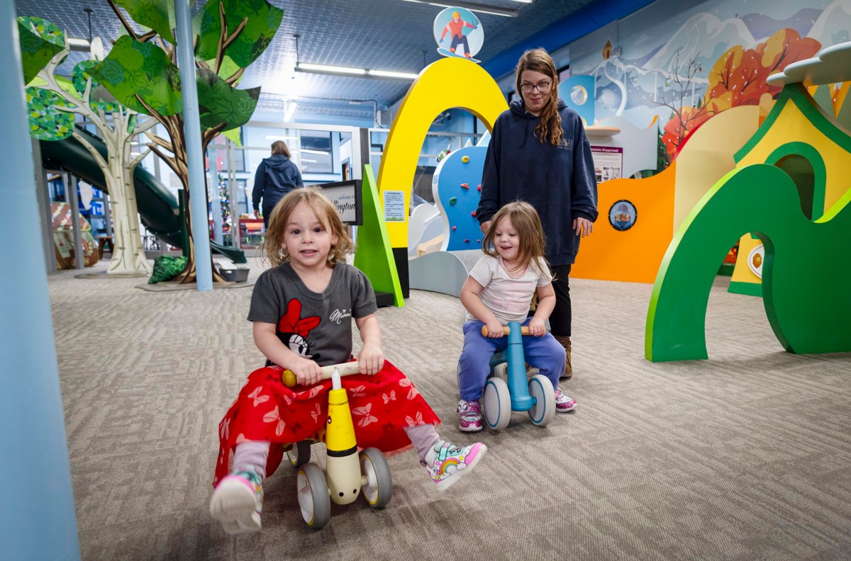 Children play on toy bikes in an indoor play area with a woman supervising. Bright and colorful structures surround them.