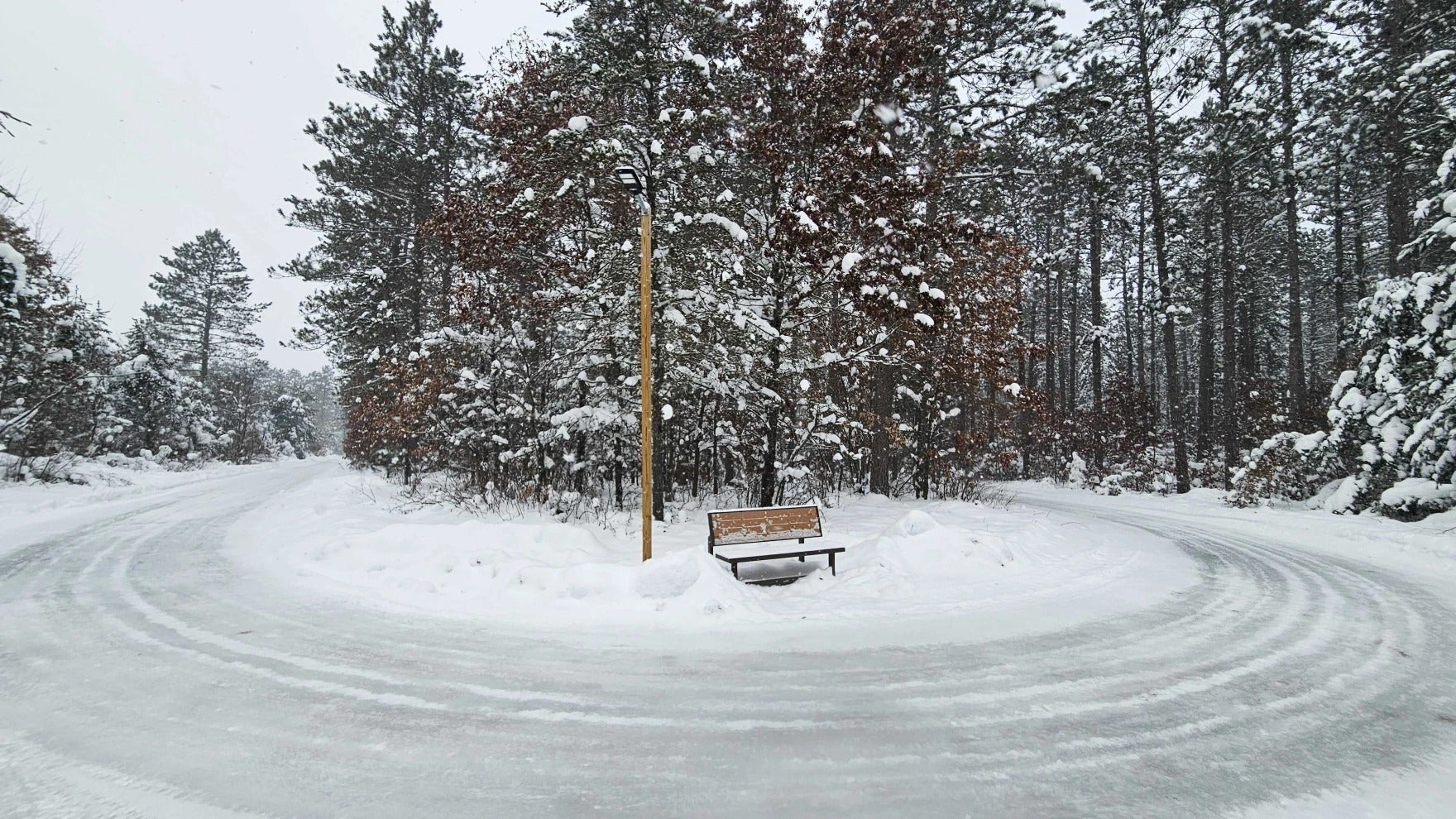A wooden bench sits on a snowy island at the center of a circular, snow-covered road lined with tall, snow-covered trees.
