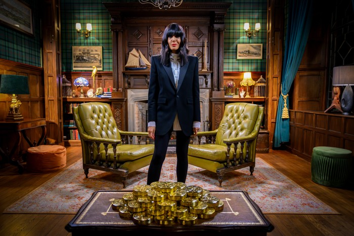 Claudia Winkleman in a tailored black suit stands confidently between two green leather armchairs in a wood-panelled room, facing a table piled high with gold bars.