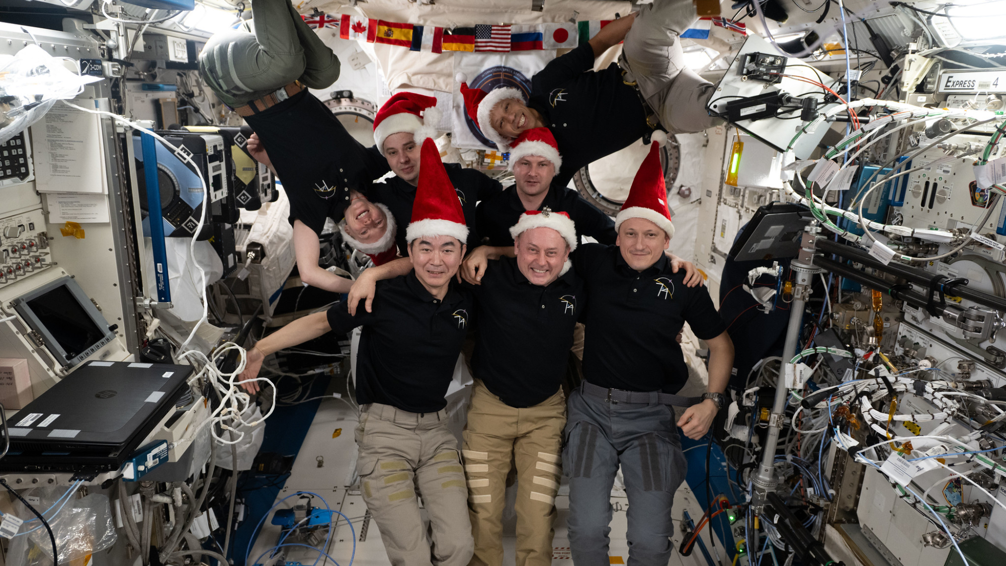 Seven astronauts wearing red and white Santa hats smiling, two are floating upside