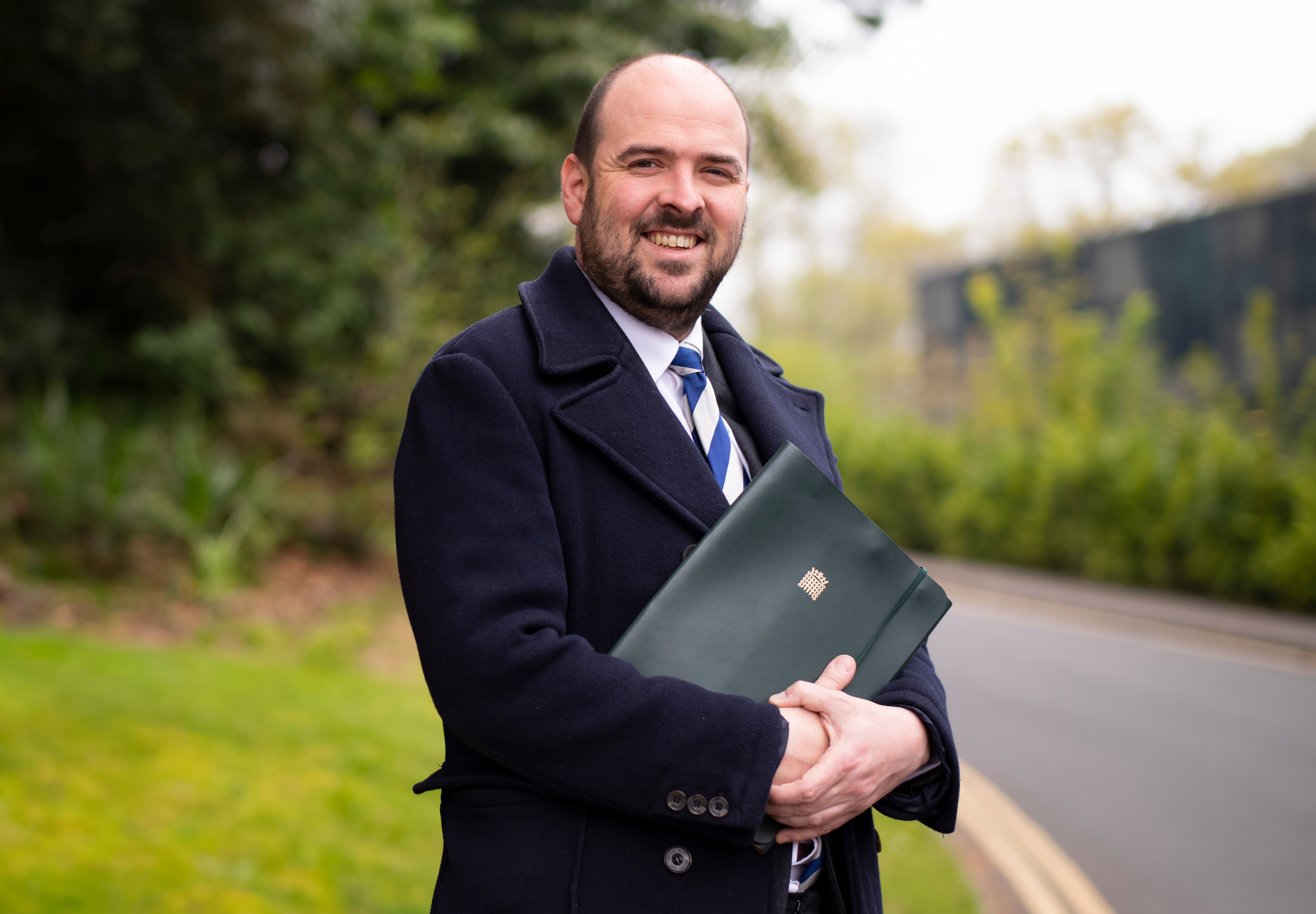 Conservative MP Richard Holden smiling and holding a dark green folder with a gold emblem.