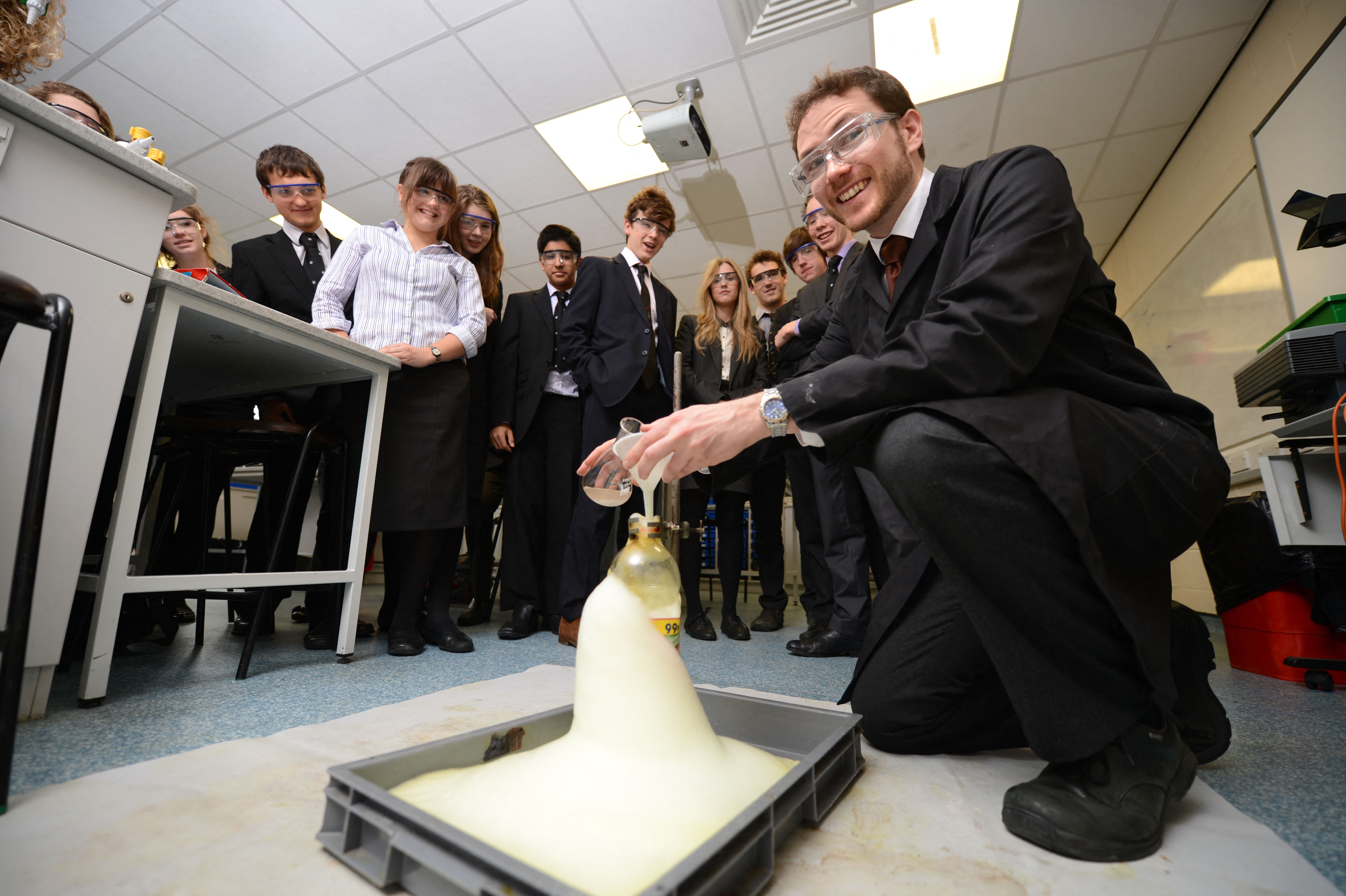 A chemistry teacher demonstrates an experiment in a classroom, with foam erupting from a bottle as students watch.