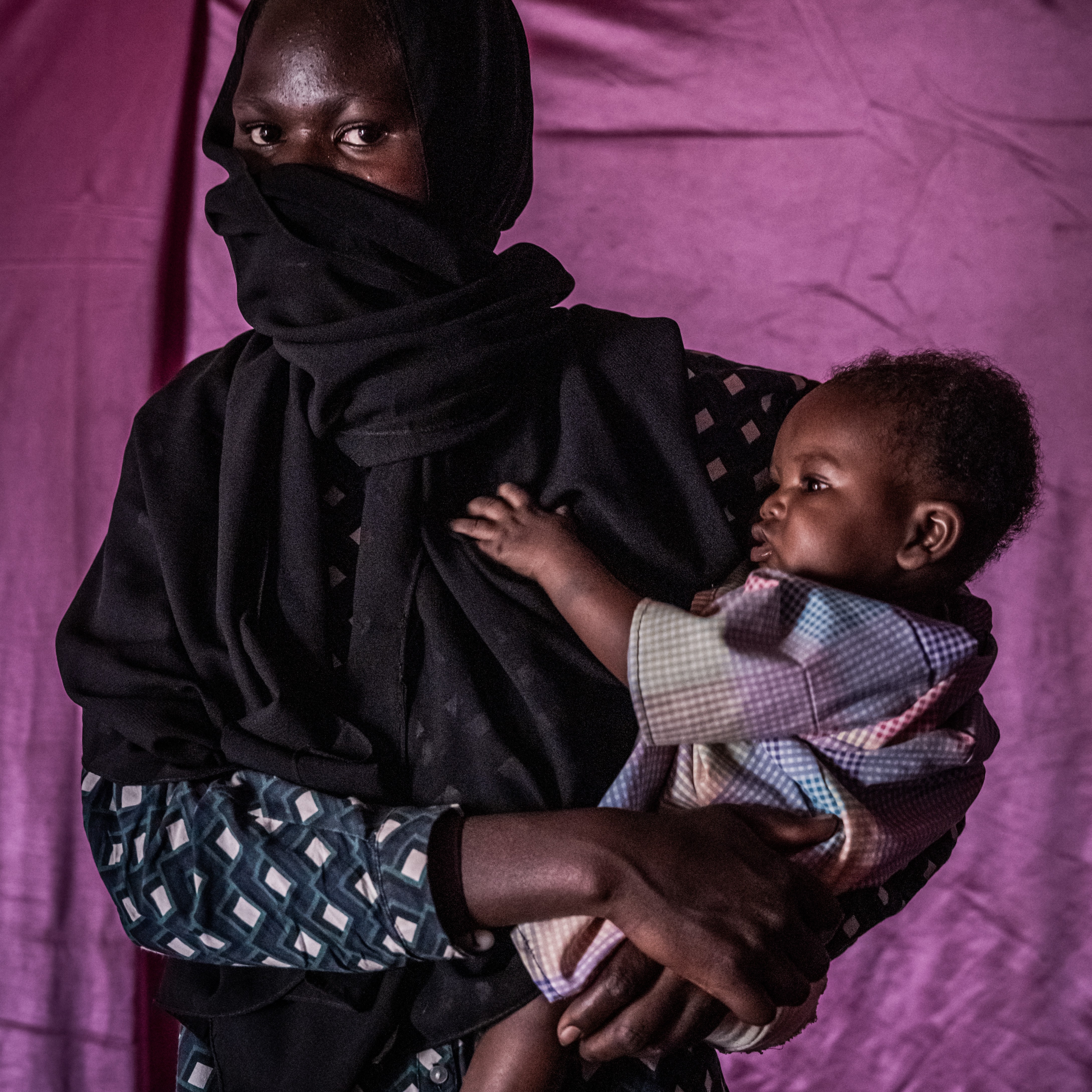 A young woman, wearing a black head covering and scarf that conceals her nose and mouth, holds a baby in her arms against a vibrant purple backdrop.