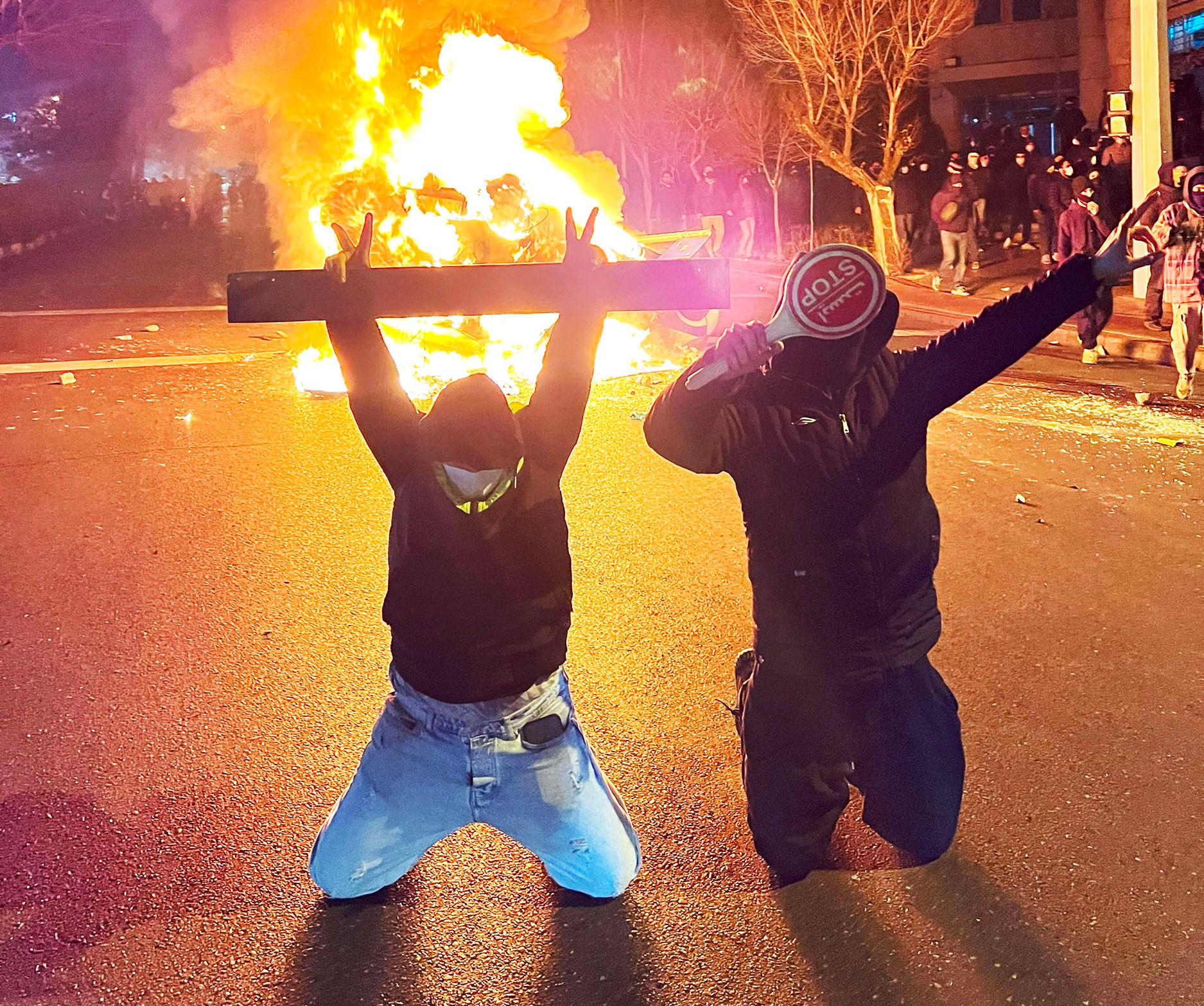 Two people kneel in front of a burning fire, one holding up a street sign and the other holding a stop sign.