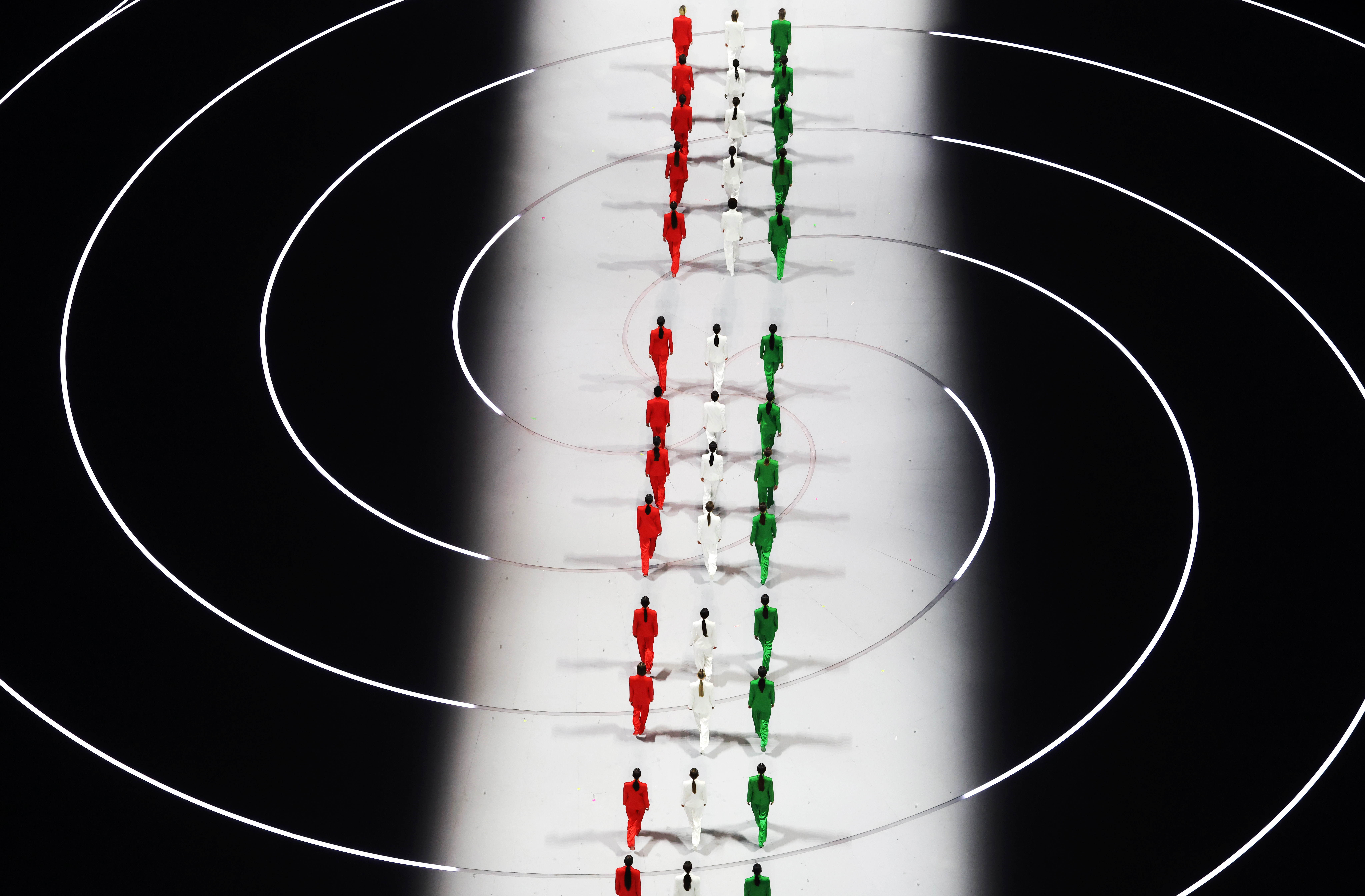 Models in red, white, and green suits walk in lines at the Milano Cortina 2026 Winter Olympics opening ceremony.