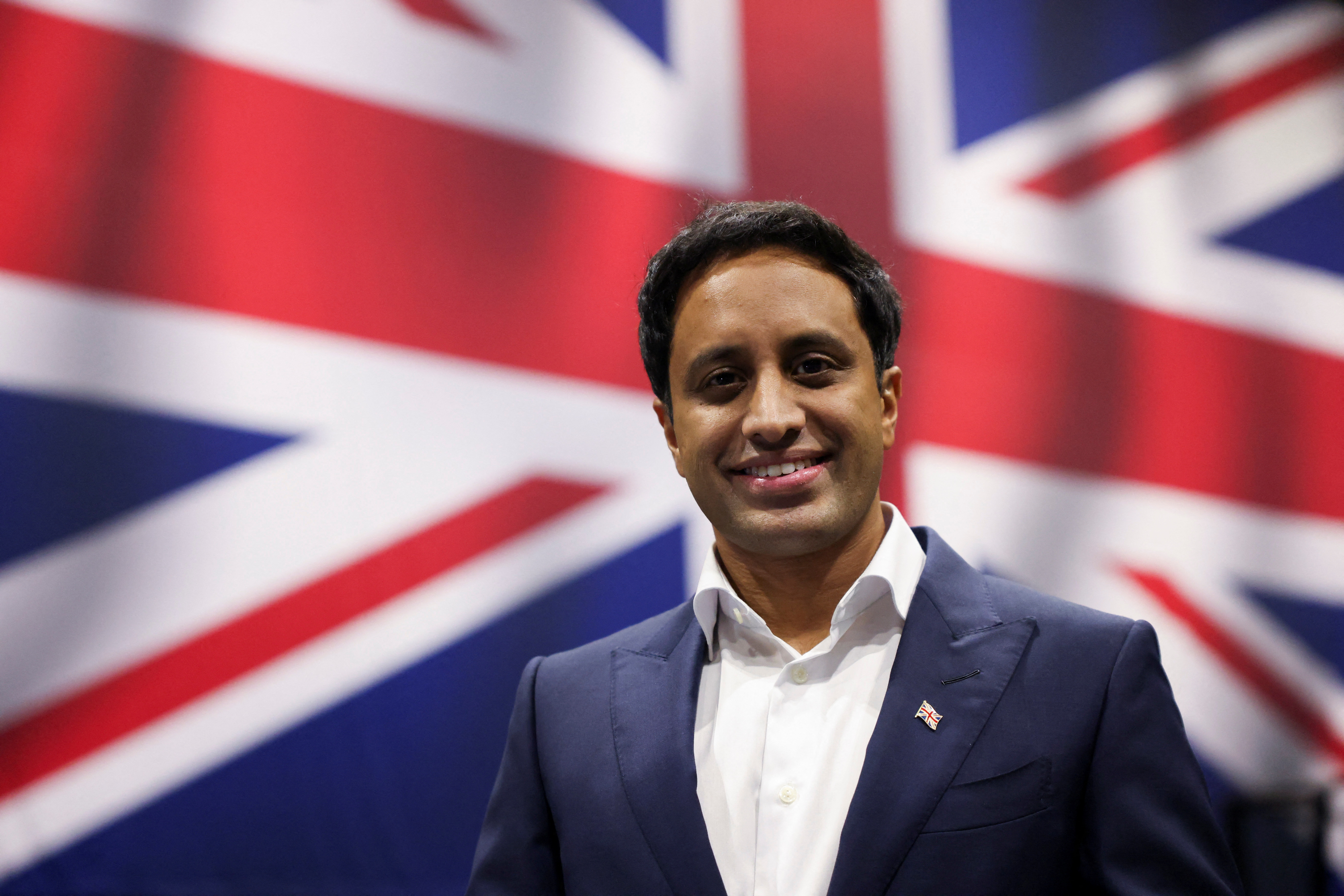 Zia Yusuf, Reform UK's largest donor, poses in a suit with a British flag pin in front of a large British flag.