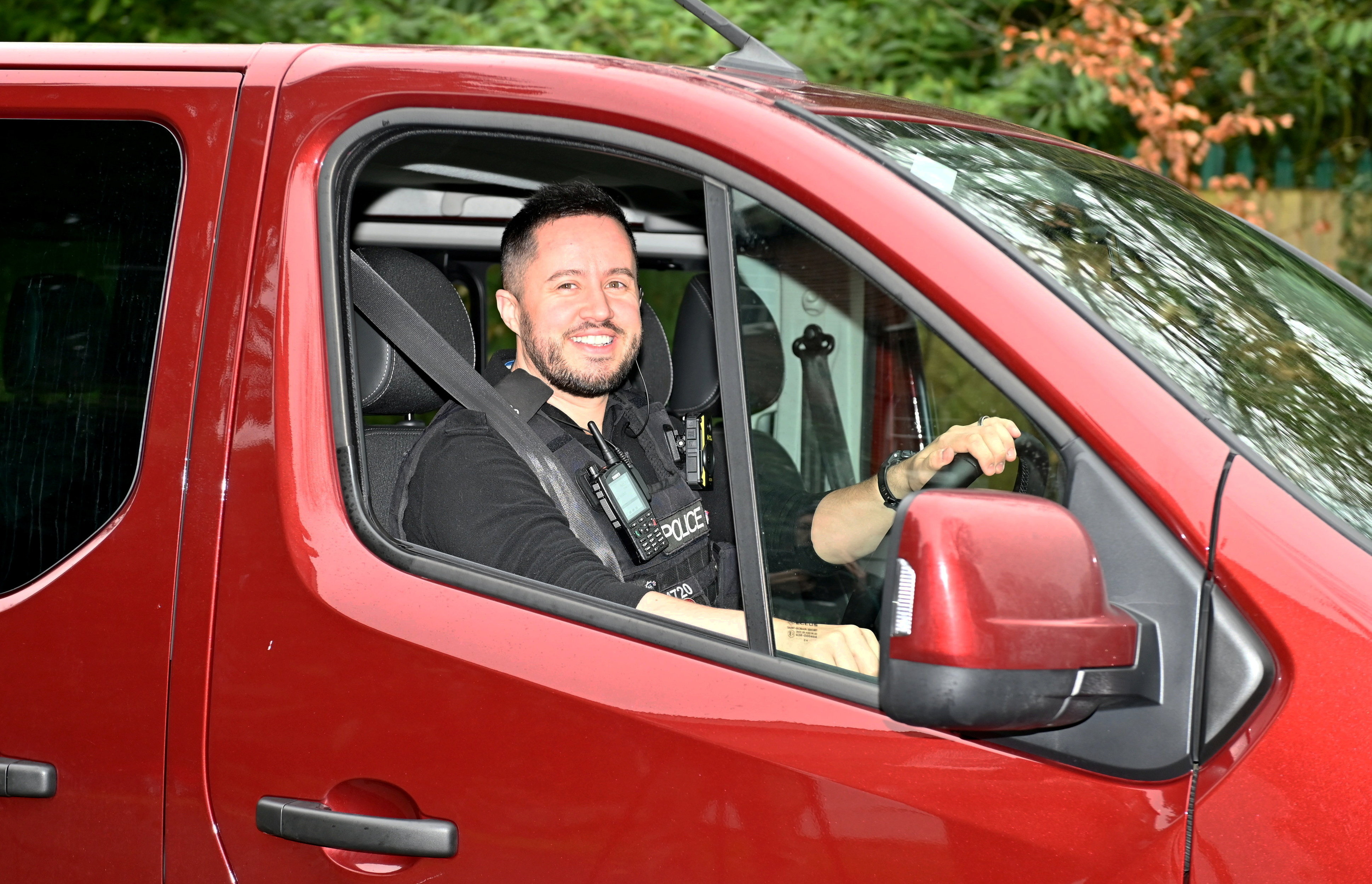 A smiling police officer in uniform and a tactical vest inside a red vehicle.