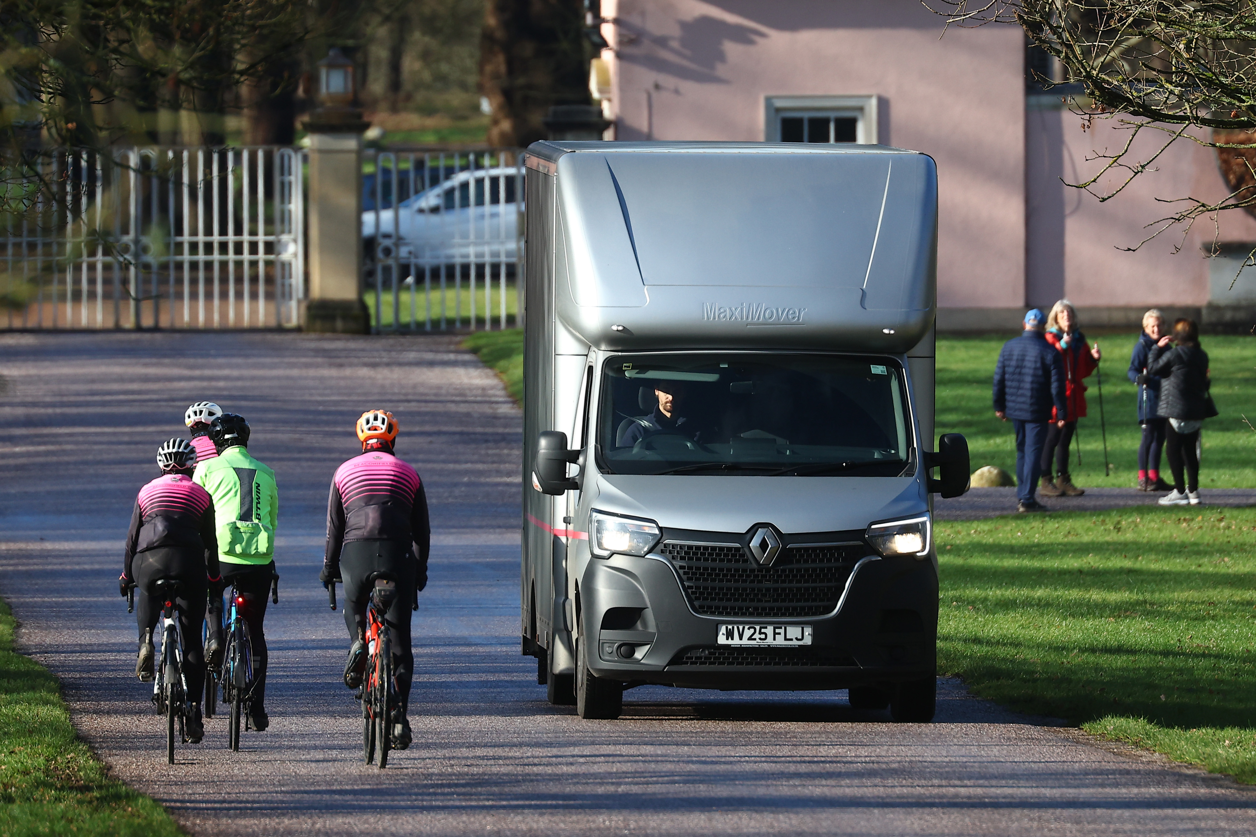 A gray removals van drives away from the gates of Royal Lodge, with three cyclists riding past it and people standing on the grass to the right.