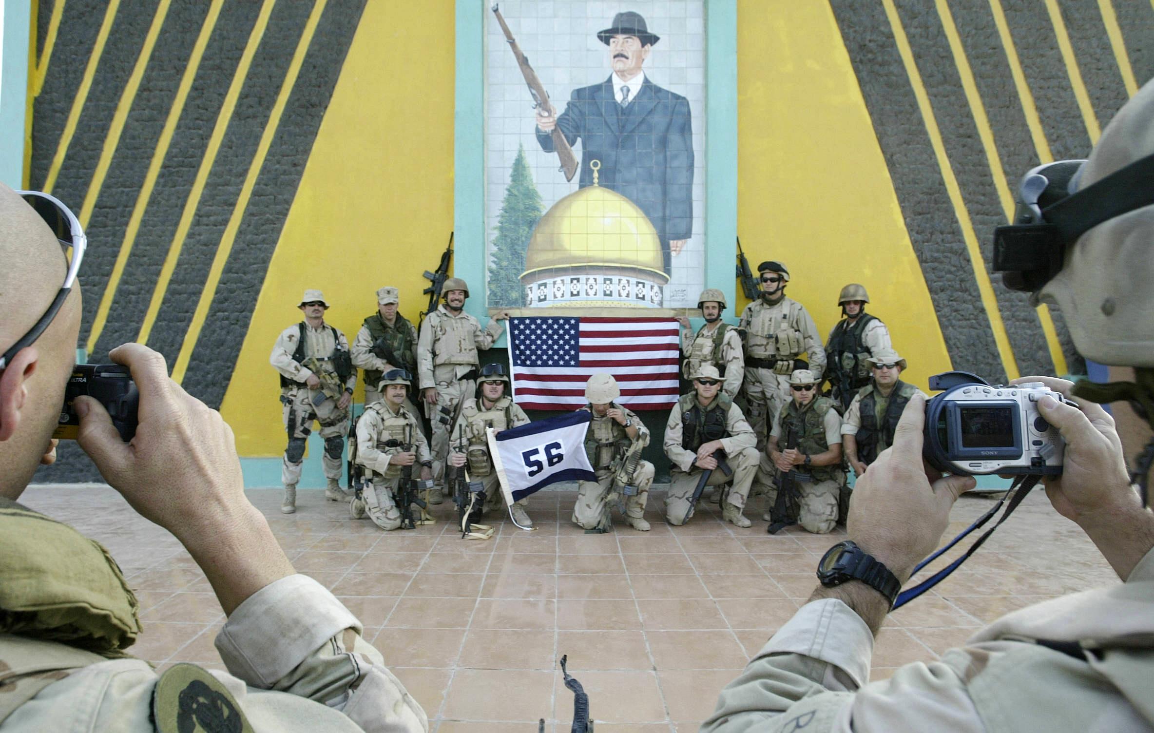 US Navy members pose in front of a picture of Iraqi President Saddam Hussein and an American flag.