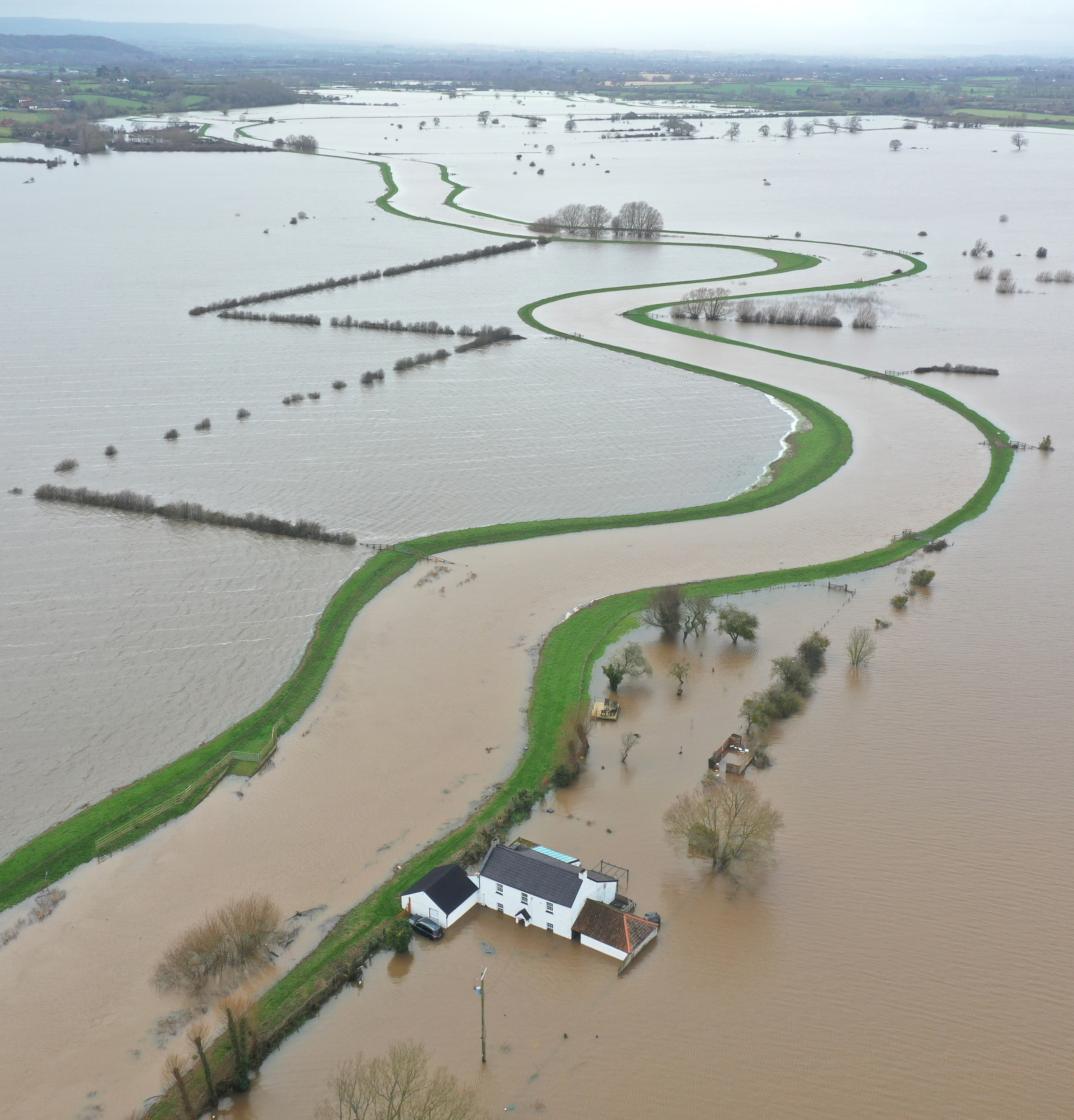 Aerial view of a white house surrounded by widespread flooding, with a winding green strip of land extending into the brown floodwaters.