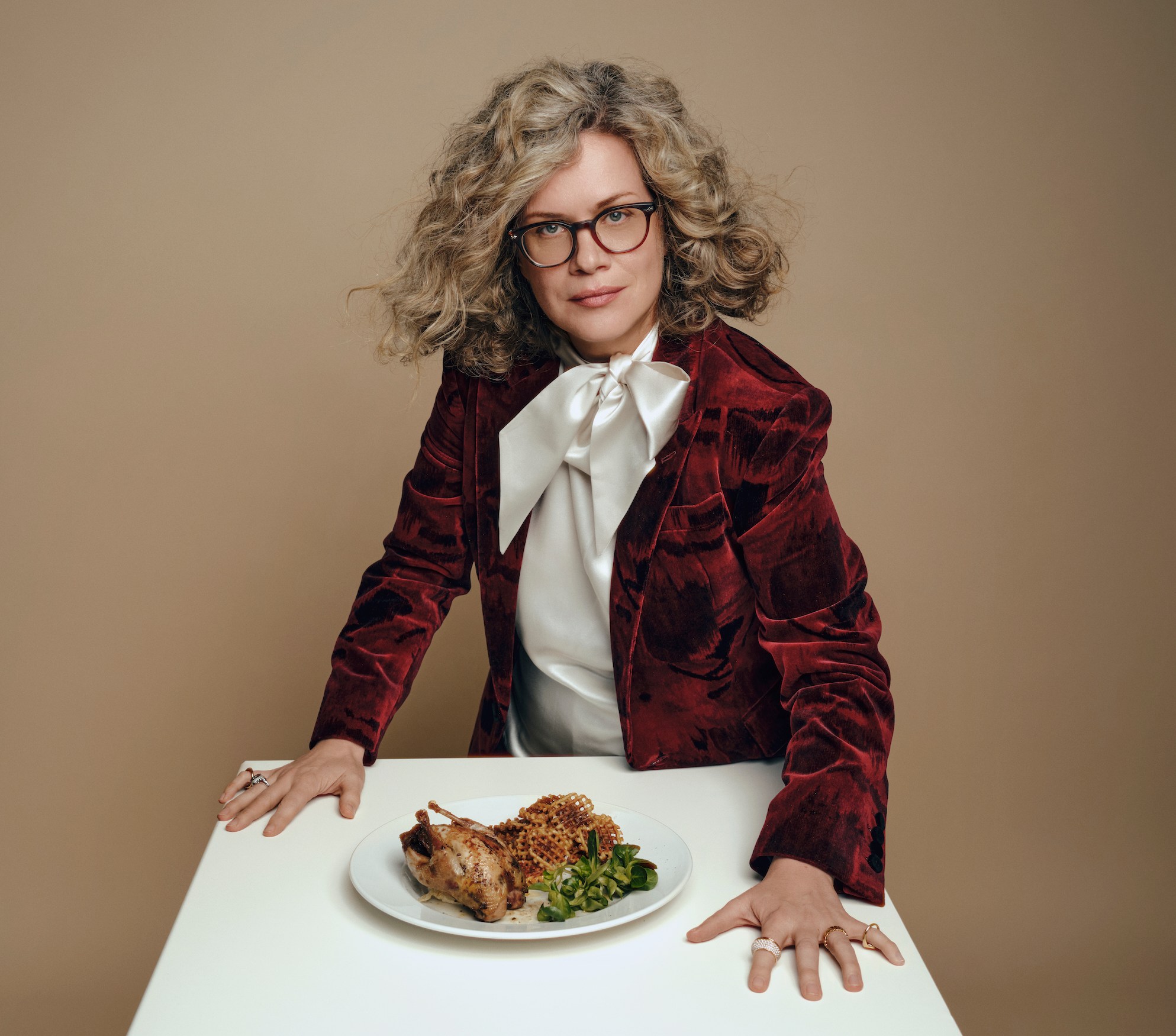 Camilla Long in a red velvet jacket and white blouse with a quail dinner on a white table.