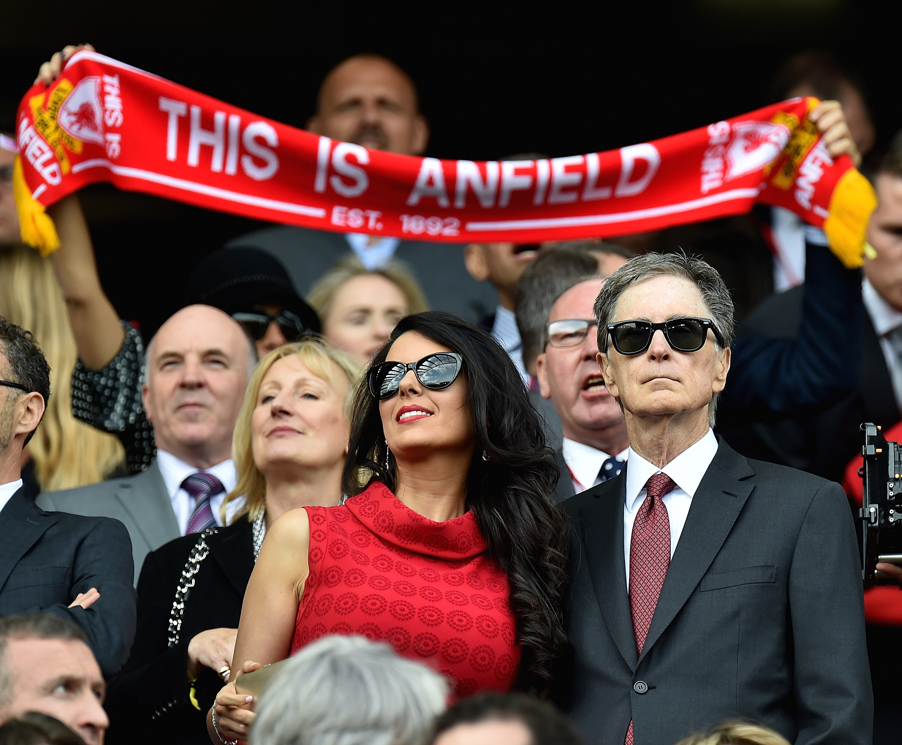 John W. Henry and his wife Linda Pizzuti in the director's box at a Liverpool FC match, with a fan holding a red "THIS IS ANFIELD" scarf in the background.