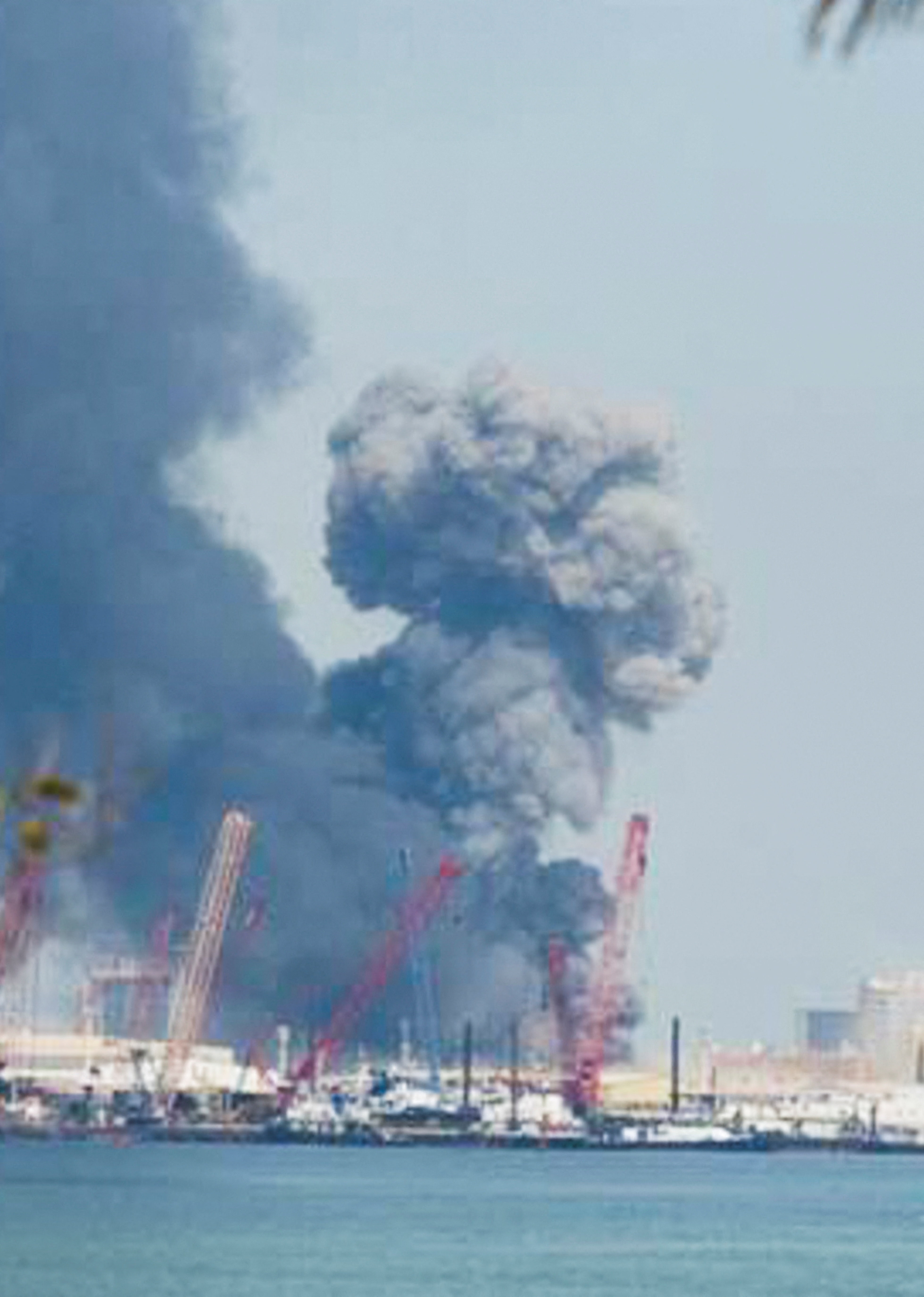 Dark smoke rises into the sky above a body of water and what appears to be an industrial area or port.