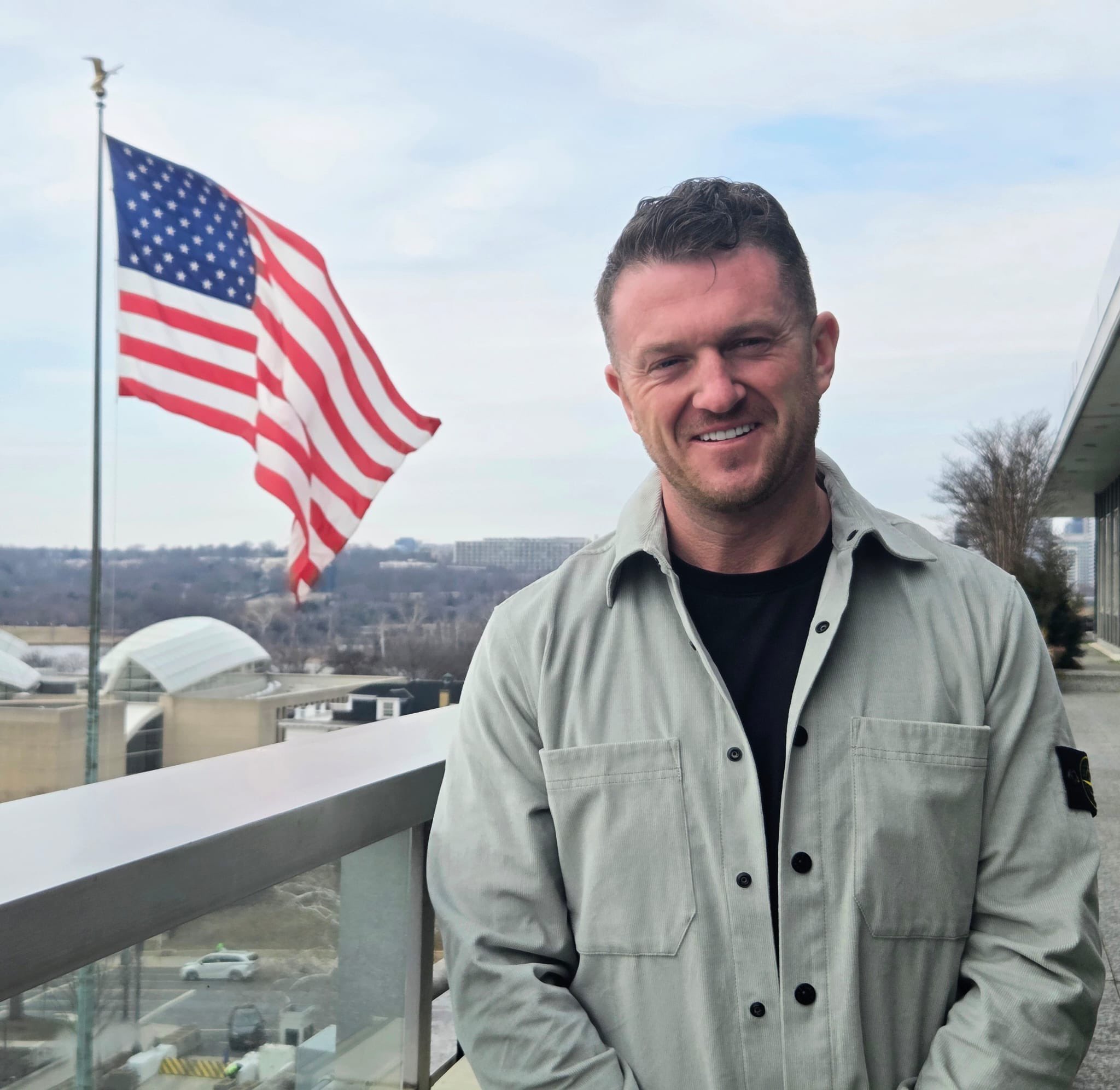 Tommy Robinson smiling with an American flag in the background, in Washington D.C.