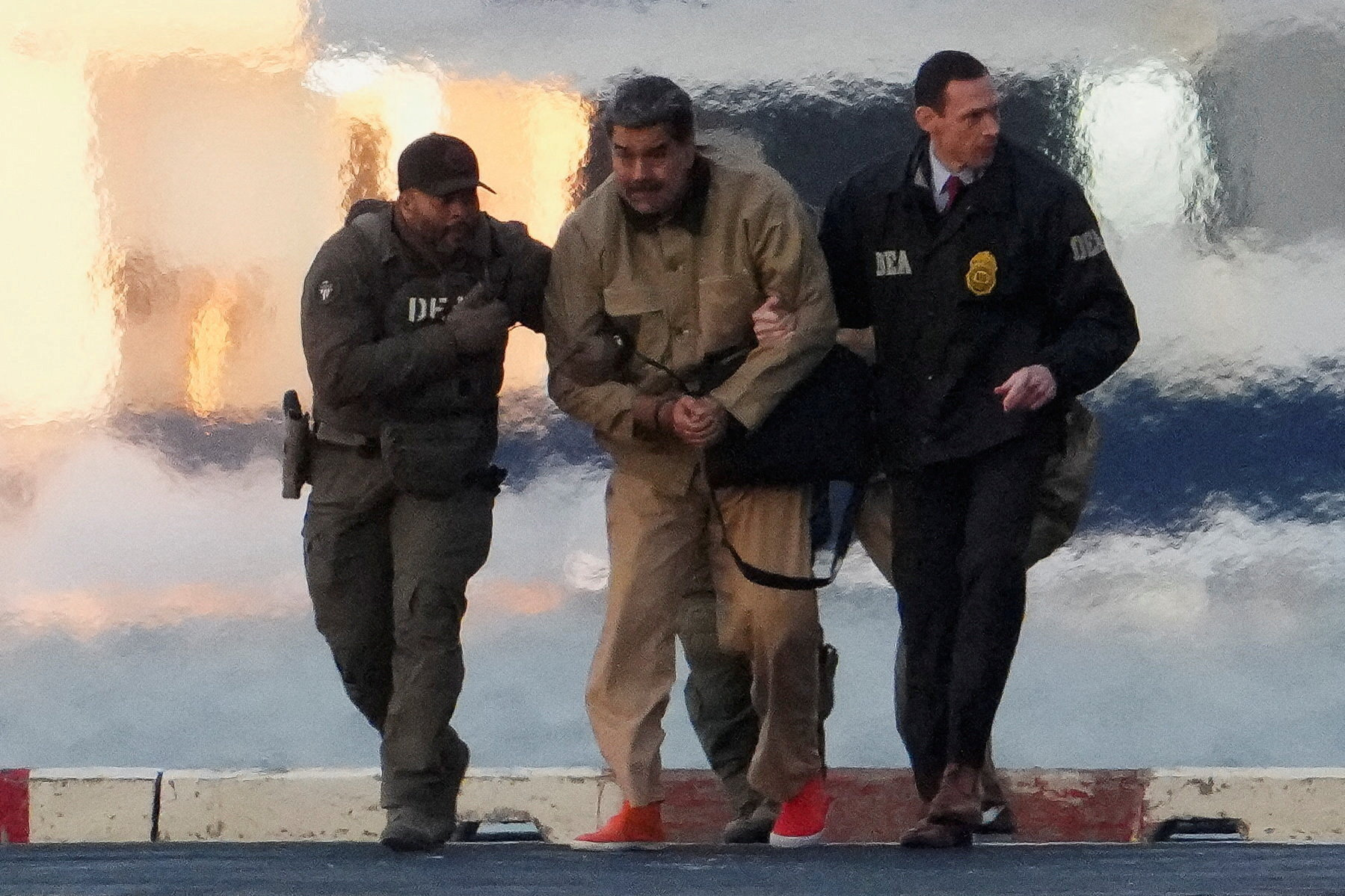 Venezuelan President Nicolas Maduro, handcuffed and wearing a tan jumpsuit and red shoes, is escorted by two men in DEA jackets.
