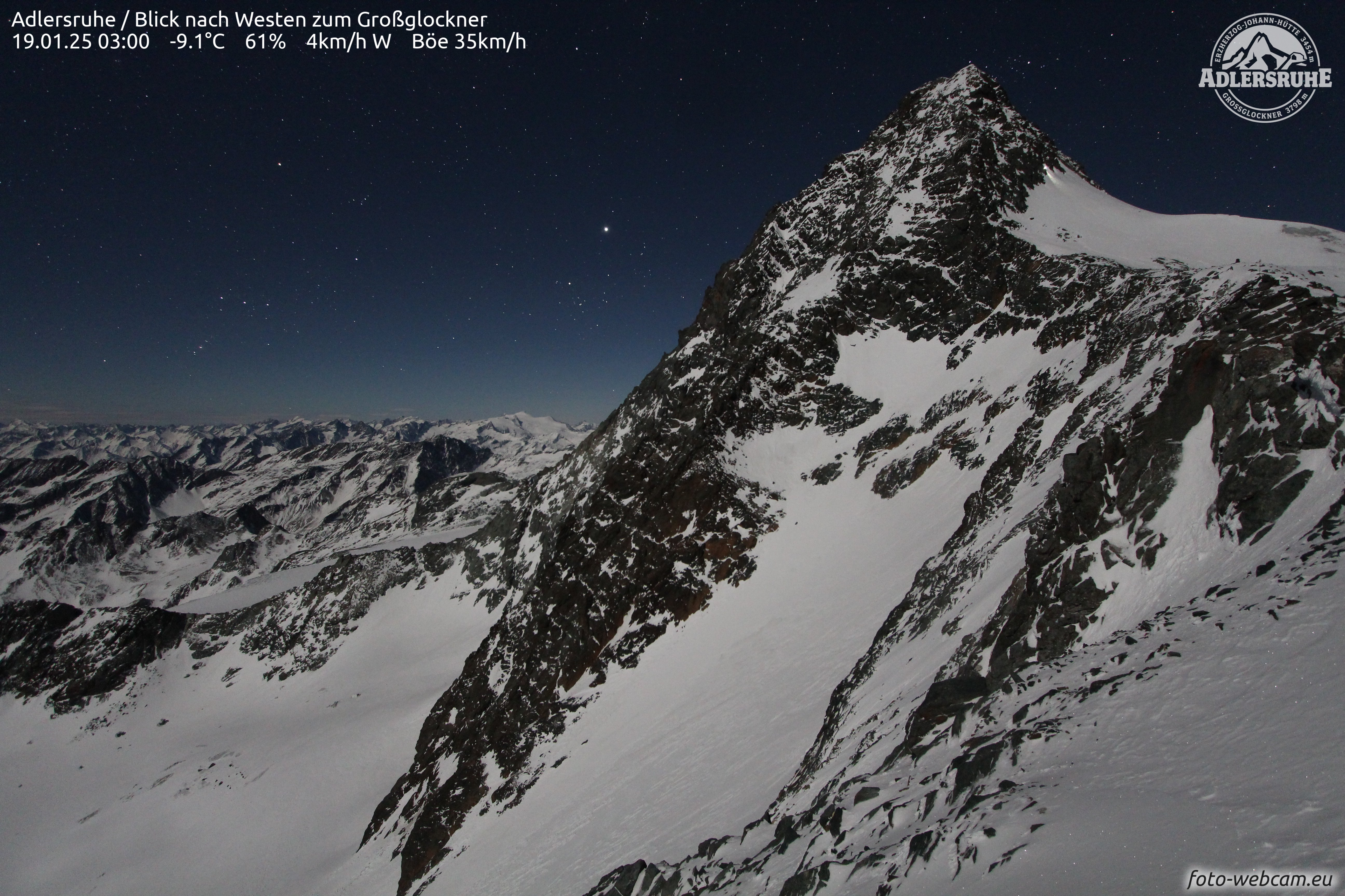Webcam footage of a boyfriend with a torch descending from the peak of Grossglockner mountain, with other snow-covered mountains in the background under a starry sky.
