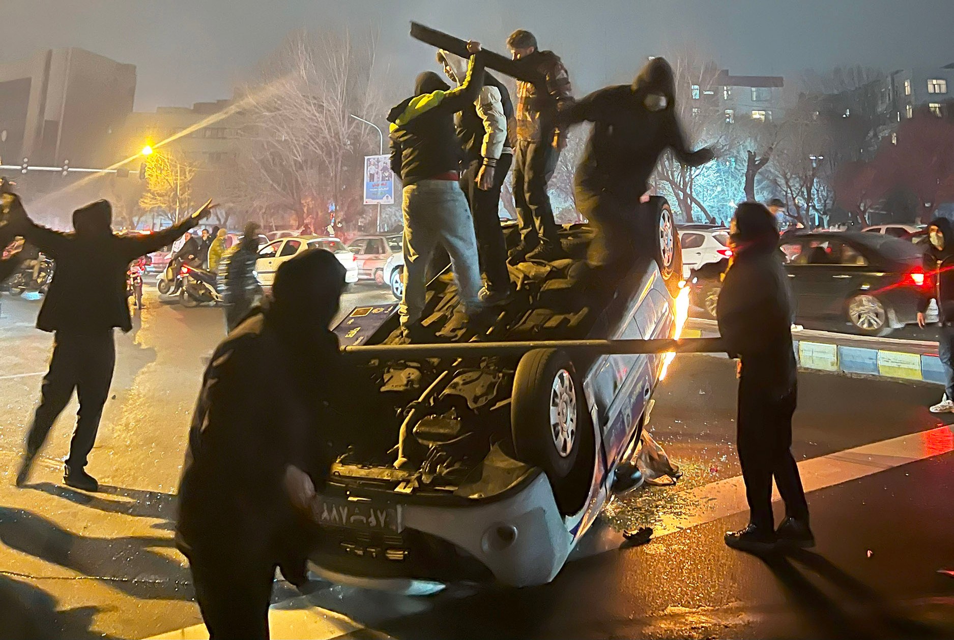 Protesters gathered around an overturned car, some standing on it, during protests in Iran.