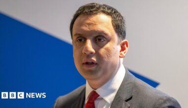 Anas Sarwar looks ahead with a serious expression on his face. He has short dark hair and wears a white shirt, red tie and black suit.