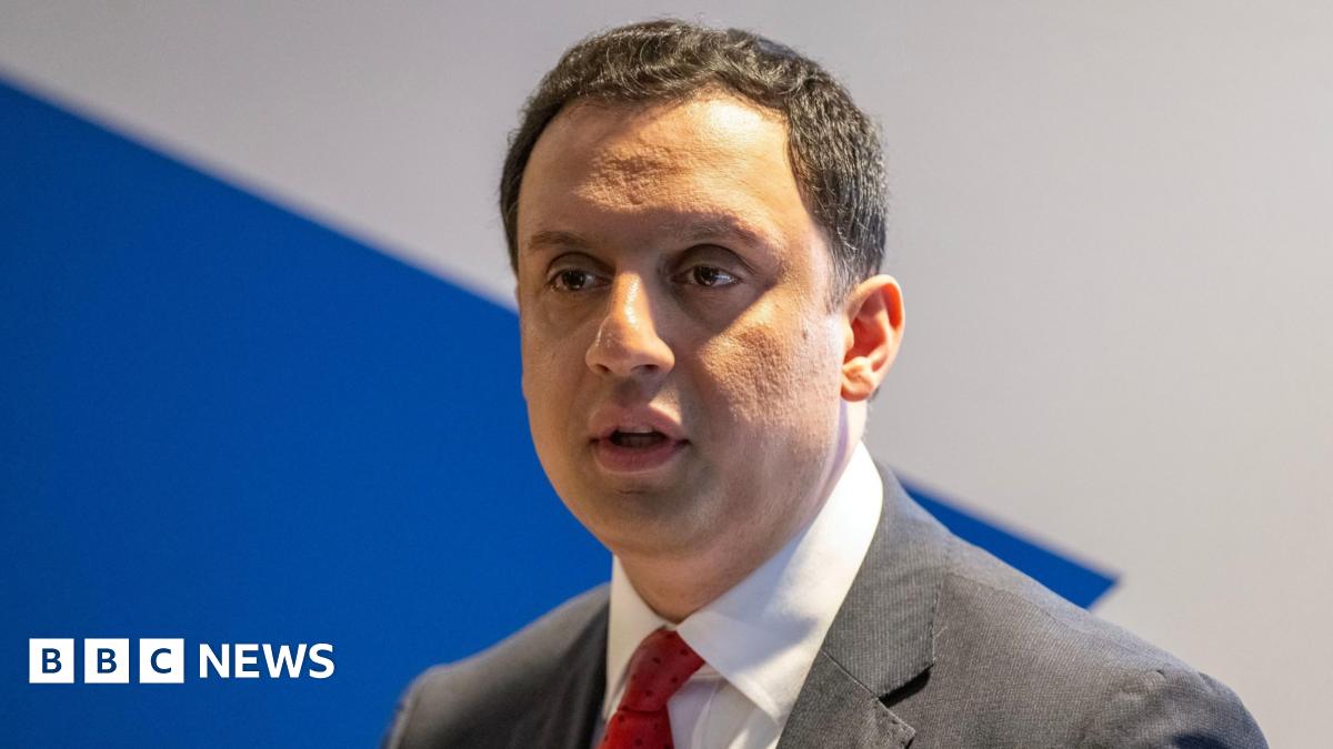 Anas Sarwar looks ahead with a serious expression on his face. He has short dark hair and wears a white shirt, red tie and black suit.
