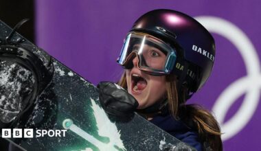 Mia Brookes shows an expression of delight, with her mouth wide open as she wears a protective dark-coloured helmet and ski goggles while carrying a snowboard