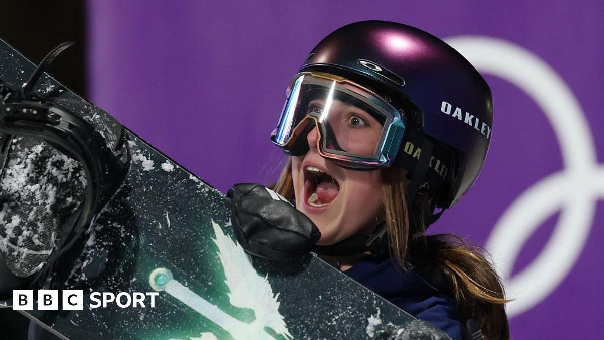 Mia Brookes shows an expression of delight, with her mouth wide open as she wears a protective dark-coloured helmet and ski goggles while carrying a snowboard