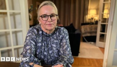 Amanda Ford sits at a table in her house. Her blonde hair is pulled back into a ponytail and she has black thick-rimmed glasses. She is wearing a blue and white paisley blouse,