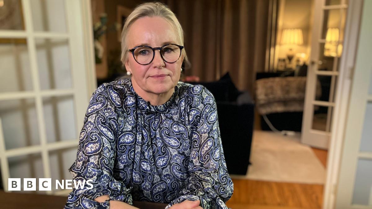 Amanda Ford sits at a table in her house. Her blonde hair is pulled back into a ponytail and she has black thick-rimmed glasses. She is wearing a blue and white paisley blouse,