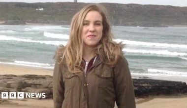 Natalie McNally, with long blonde hair, wearing a dark green coat. She is standing on a beach with the sea visible behind her.