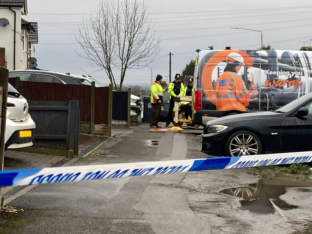 Emergency services personnel are gathered around a van and a car, cordoned off with blue and white tape, in a residential area.