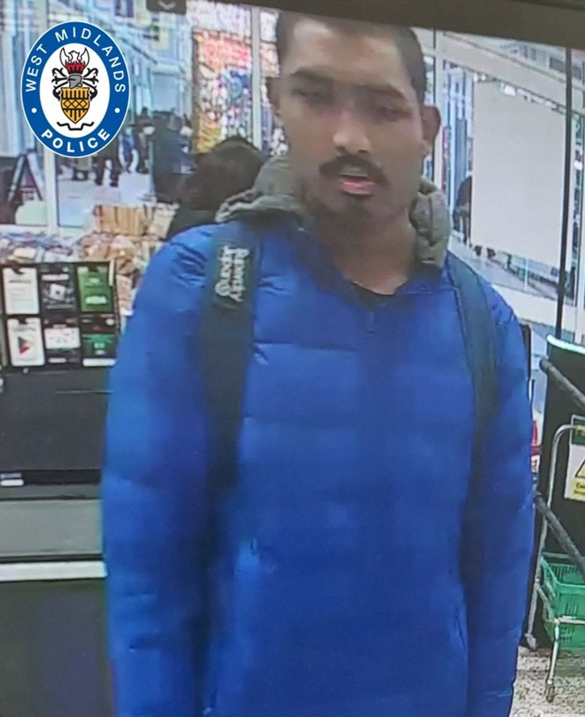 CCTV image of a man in a shop who is of Asian appearance with short black hair, a moustache and goatee beard, pictured wearing a royal blue puffer jacket, over a hooded top, and is wearing a rucksack, grey jogging bottoms and black shoes