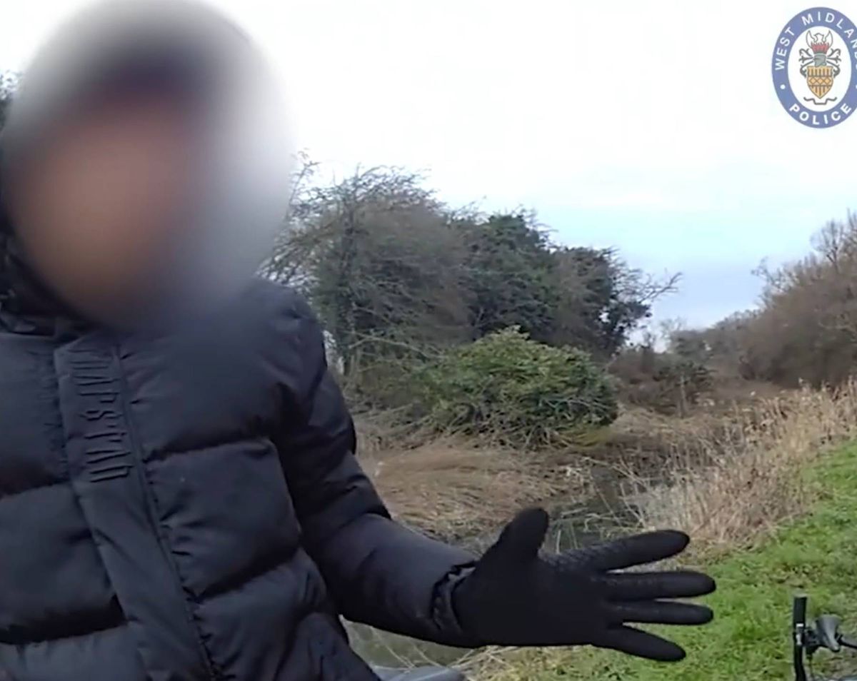 An individual dressed in a black puffy jacket and black gloves stands outdoors in a grassy area with a blurred background, appearing to gesture or point. The backdrop includes sparse vegetation and a distant horizon.