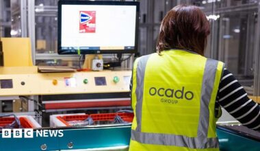 A worker in an Ocado distribution centre wearing a hi-vis jacket with Ocado Group written on the back, The worker is looking at a screen and filling baskets