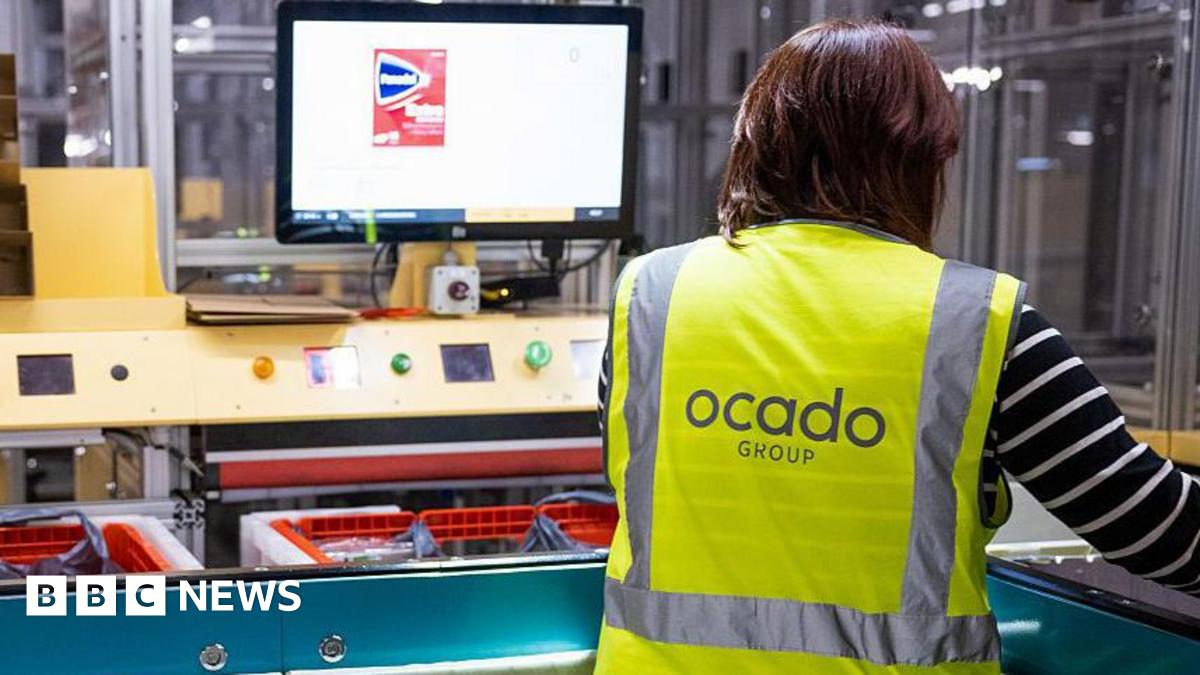 A worker in an Ocado distribution centre wearing a hi-vis jacket with Ocado Group written on the back, The worker is looking at a screen and filling baskets