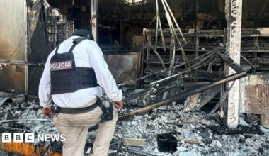 An image showing muicipal police trucks next to a burning bus set on fire, and a police man is walking in between. The bus is charred with flames engulfing it from below. The cars are parked on a highway.