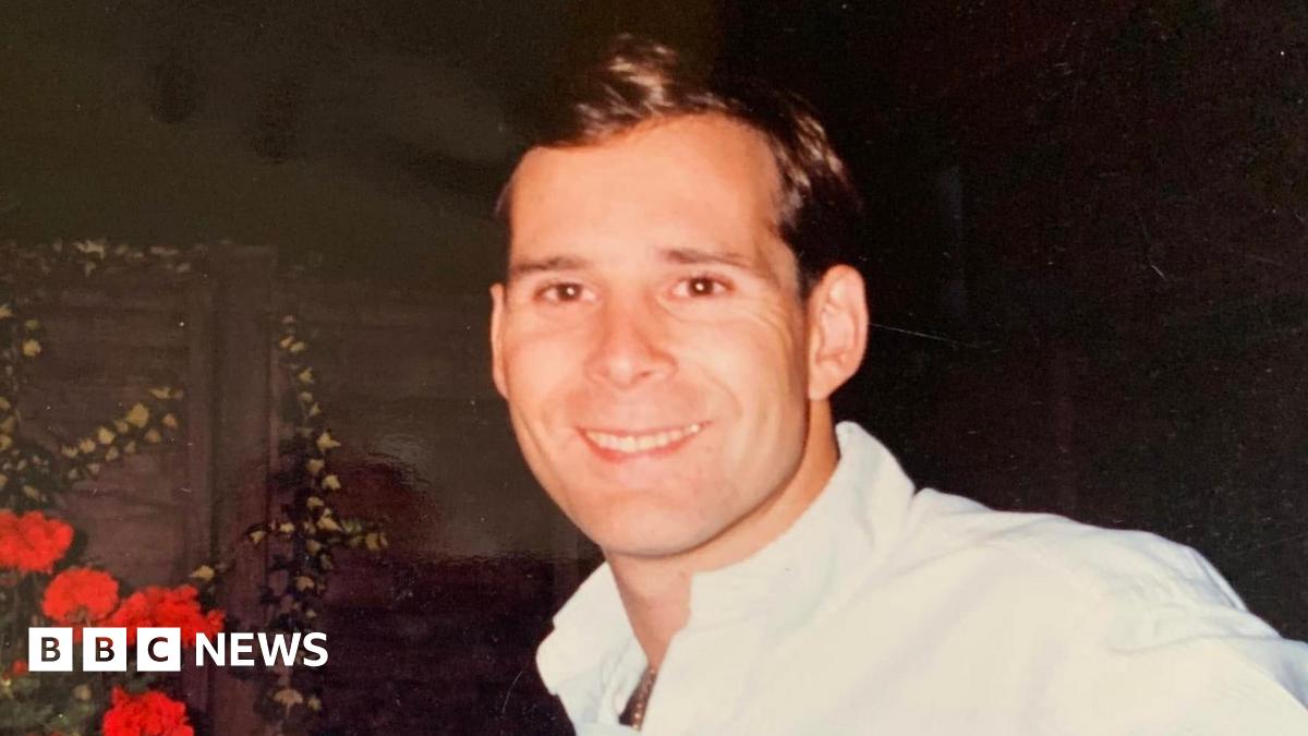 A photograph of a man stood in a garden next to red flowers wearing a white shirt. He is clean shaven, with short dark hair.
