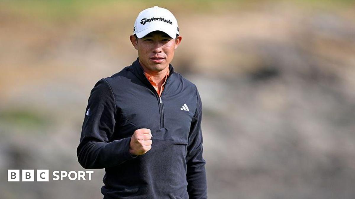 Collin Morikawa clenches his right fist after sinking his winning putt. He is wearing a white cap and black zipped-up top.