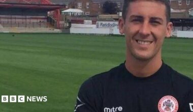 A man in a football kit for Accrington Stanley standing on a pitch, smiling. He has brown hair.