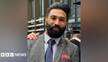 A man with black hair and a beard smiles at the camera, wearing a white and blue checked shirt, blue tie with small red spots, and a grey tweed jacket.