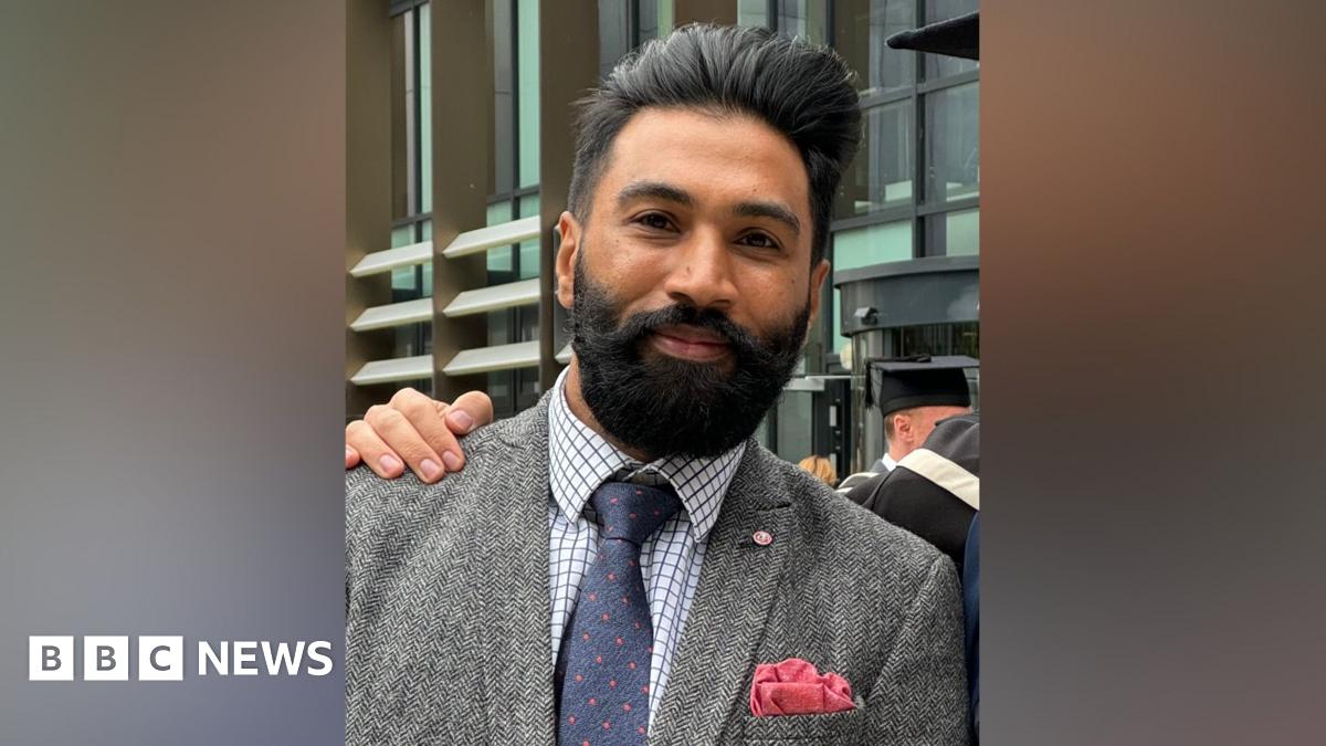 A man with black hair and a beard smiles at the camera, wearing a white and blue checked shirt, blue tie with small red spots, and a grey tweed jacket.
